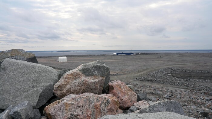 De grosses roches empilées, avec en arrière-plan un bâtiment de la mine et un plan d'eau, en août 2024.