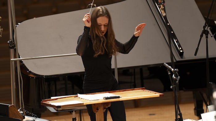 Dina Gilbert, debout sur un podium avec une baguette, dirige des musiciens.