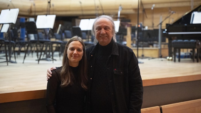 Dina Gilbert et Claude Dubois sourient côte à côte devant une scène d'orchestre avec des lutrins et un piano à queue.