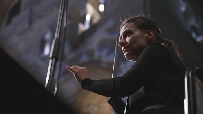 Vue en contre-plongée d'une cheffe d'orchestre pointant sa baguette, dans une basilique. 