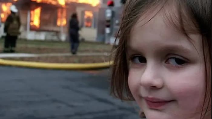 Une petite fille regarde l'objectif de la caméra avec un air coquin alors qu'un feu ravage une maison derrière elle.