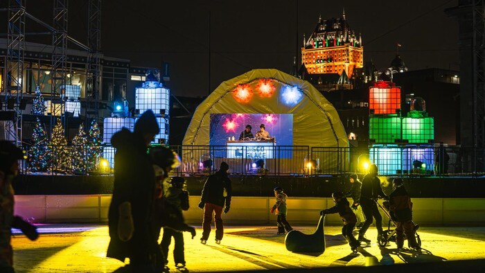 Une vue nocturne animée de la Discoglace au Port de Québec, où des patineurs évoluent sur une glace illuminée de jaune devant un DJ installé sous un dôme gonflable, avec le Château Frontenac éclairé qui surplombe la scène à l'arrière-plan.