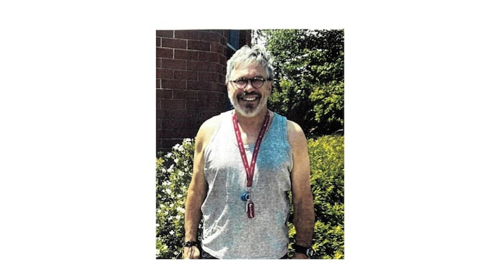 Un homme avec des lunettes rondes, des cheveux courts et une barbe, pose devant un buisson. La photo a été prise en été.