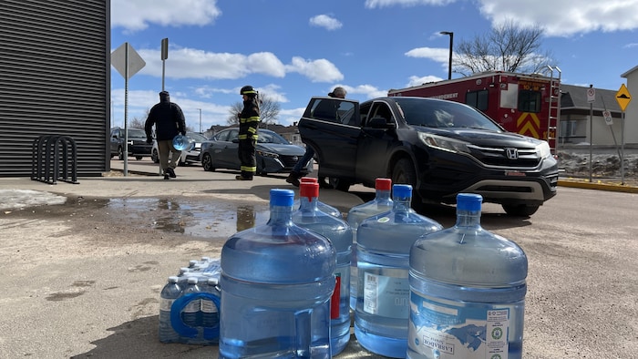 Six bouteilles de 18 litres et un paquet de petites bouteilles sont posées par terre dans un stationnement. 