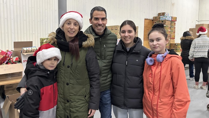 Une famille de quatre personnes, tous vêtus de manteaux d'hiver, sourit pour une photo. La mère et le jeune fils portent des chapeaux de père Noël. Ils sont dans un entrepôt où des boîtes cadeaux emballées sont visibles à l'arrière-plan.