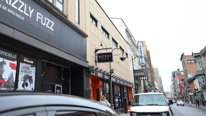 Vue en contre-plongée de la façade de la salle de spectacle Grizzly Fuzz dans une rue de Québec. L'enseigne principale indique "GRIZZLY FUZZ SALLE DE SPECTACLE". Des affiches d'événements sont visibles à l'avant, dont une pour "LOUD LARY AJUST". La rue est bordée d'autres commerces.