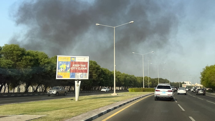 Des voitures circulent sur une route. Derrière, au loin, de la fumée s'élève dans le ciel.
