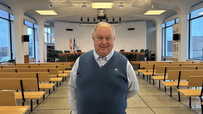 Un homme sourit à la caméra, au centre d'une salle de conseil municipal vide.