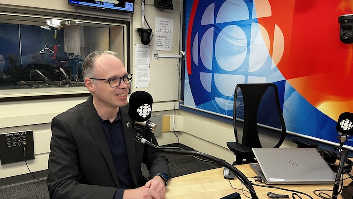 un homme avec des lunettes et un veston assis au micro dans un studio de radio de Radio-Canada