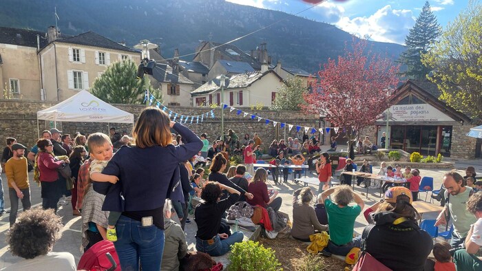 Une foule au beau soleil sur une place publique d'un village situé au pied d'une montagne.