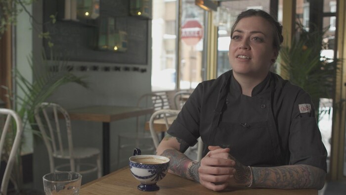On voit Mme Dufour assise à une table de son restaurant, en train de parler à la caméra.