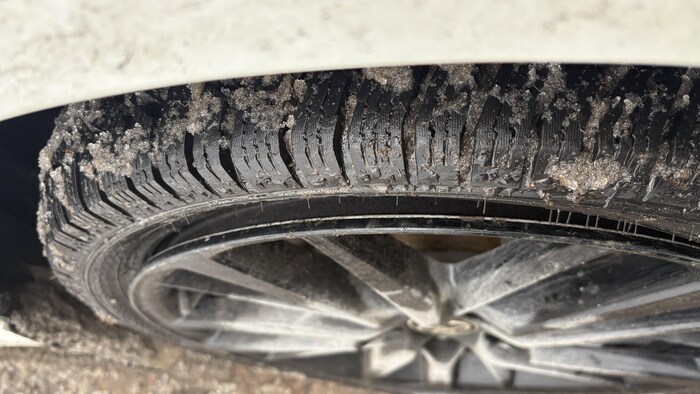 Photographie en gros plan d'un pneu de voiture noir aux rainures profondes, partiellement couvert de neige fondue et de boue grise, monté sur une jante à rayons multiples. Le pneu est positionné sous l'aile blanche d'un véhicule, sur une chaussée mouillée.