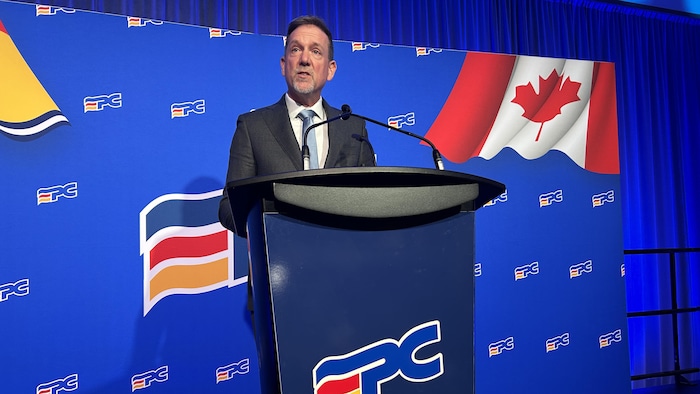 Don Monahan au lutrin, sur un fond bleu, parle en jetant un regard vers la gauche.