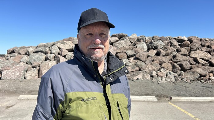 Un homme portant un coupe-vent et une casquette est debout devant un mur de grosses roches haut d'environ deux mètres.