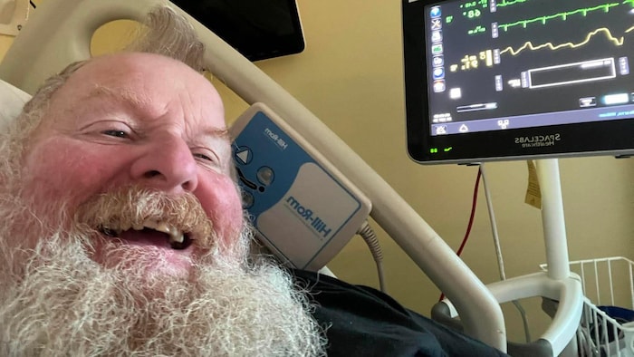 Un homme sur un lit d'hôpital pose souriant avec des machines autour de lui.