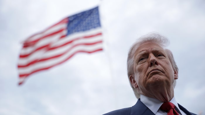 Donald Trump, debout dehors, l'air sérieux, devant un drapeau des États-Unis en berne.