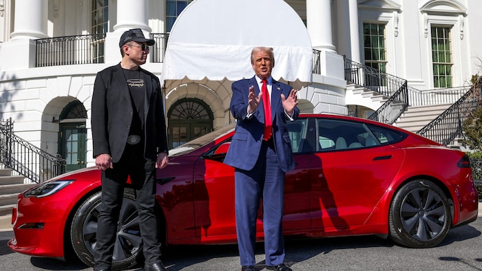 Debout devant une voiture sport Tesla rouge stationnée devant la Maison-Blanche, Donald Trump fait un geste avec ses mains. Elon Musk est debout à sa droite et le regarde.