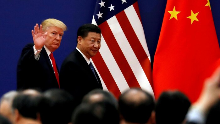 Donald Trump, qui lève la main en guise de salutation, et Xi Jinping marchent sur une scène devant une foule. Derrière sont accrochés les drapeaux des États-Unis et de la Chine.