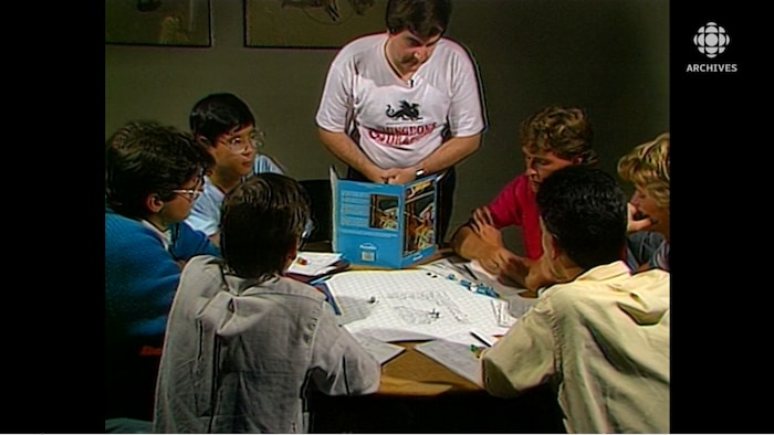 Joueurs autour d'une table, feuilles de jeu au centre et maître du jeu debout derrière son livre de scénarios. 