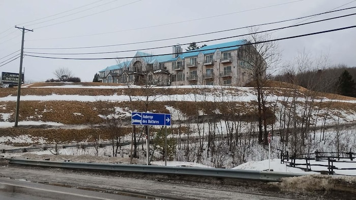 Cette vue extérieure présente l'Auberge des Battures située sur une colline enneigée. L'édifice possède une toiture bleue distinctive et un revêtement en bois clair. Au premier plan, un panneau de signalisation bleu indique la direction de l'établissement.