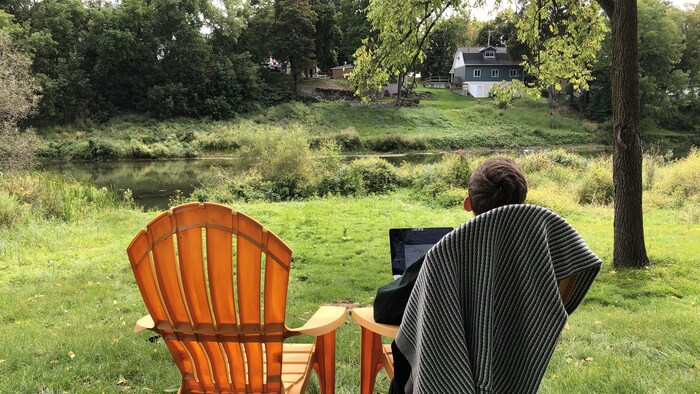 Un homme assis sur une chaise Muskoka avec son ordinateur portable, devant une rivière.