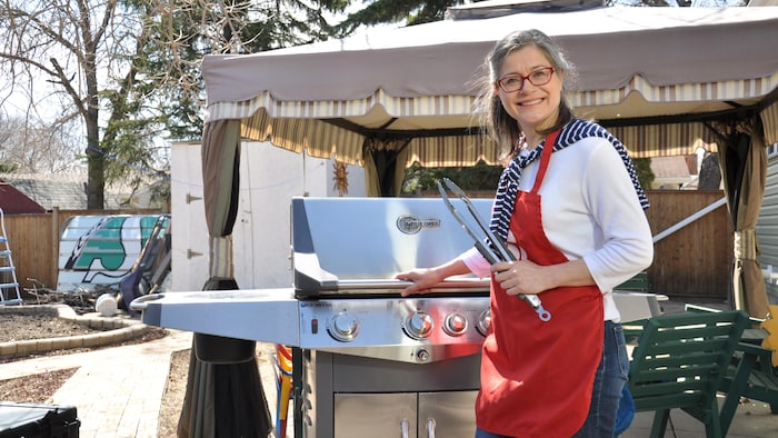 Doris Labrie est debout devant un barbecue à l'extérieur, tenant des ustensiles de cuisine. Elle porte un tablier rouge, un haut à manches longues blanc. Elle sourit en regardant vers l'appareil photo.