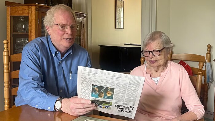 Deux personnes âgées assises à une table lisent un journal en papier.