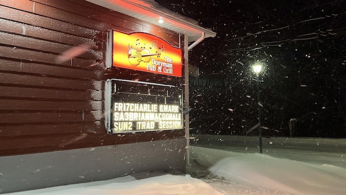 Le mur de côté du Doryman Pub, avec son enseigne, un tableau éclairé qui annonce les prochains musiciens qui vont jouer, photographié le soir pendant une tempête de neige.