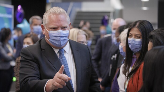 Doug Ford, portant un masque, pointant du doigt, entouré de professionnels de la santé.