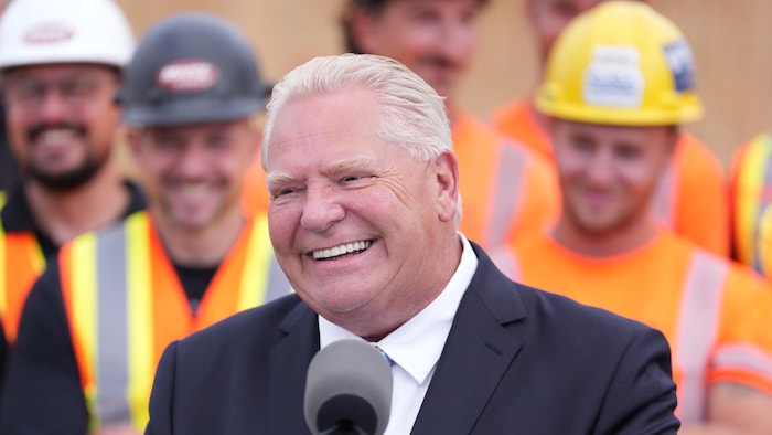 Doug Ford parlant à un lutrin devant des travailleurs de la construction portant gilet orange et casque sur un chantier de Hamilton le 24 août 2026.