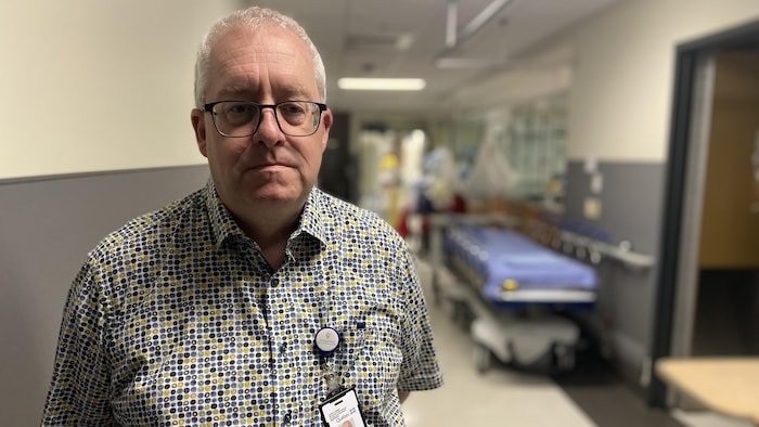 Un homme debout dans un couloir d'hôpital regarde la caméra le temps d'une photo. Il porte des lunettes.