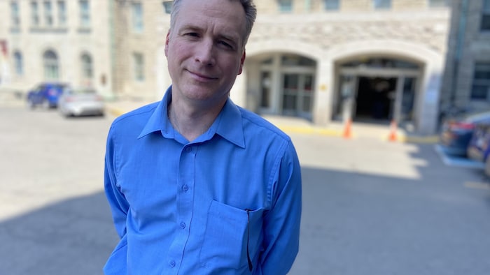 Un homme avec une chemise bleue regarde la caméra devant un hôpital.