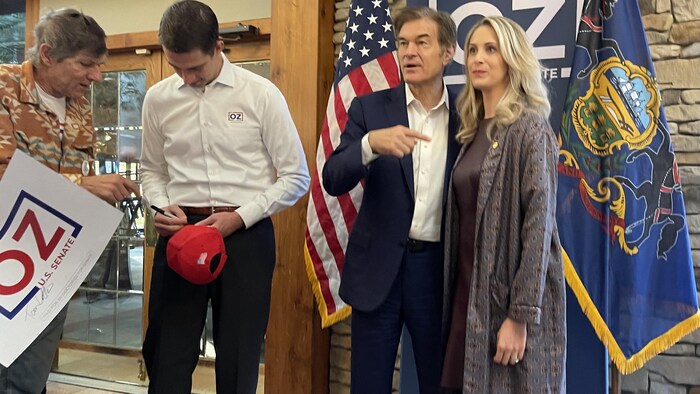 Mehmet Oz se fait prendre en photo avec une femme. À ses côtés, un homme signe une casquette « Trump, Save America ».