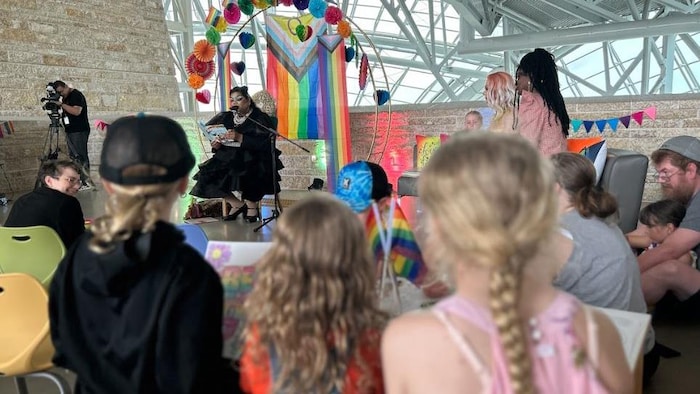 Des enfants sont debout et écoutent une drag queen au loin.