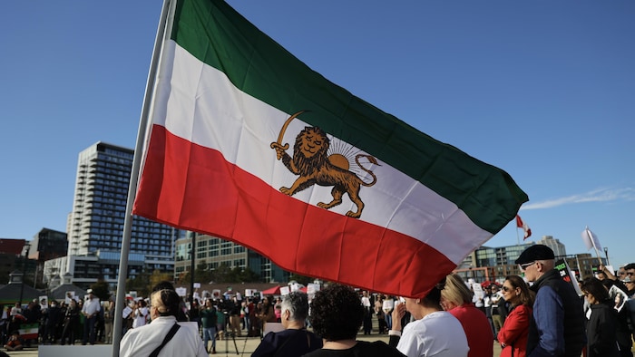 Un drapeau à barres horizontales verte, blanche et rouge avec un emblème composé d'un lion et d'un soleil au milieu flotte sur une place publique où des gens sont rassemblés pour une manifestation.