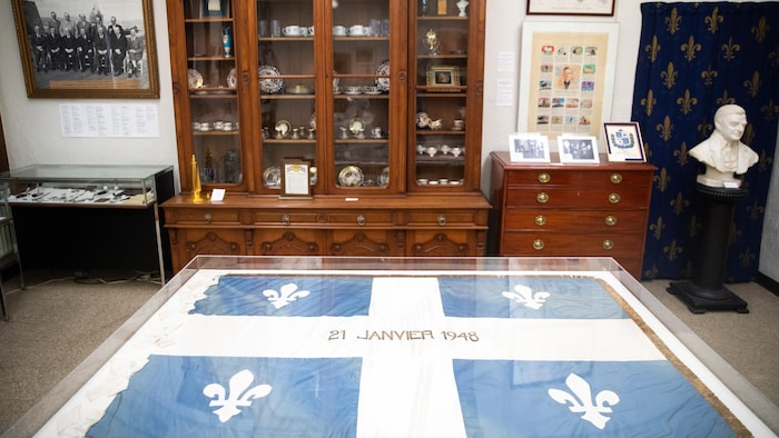 Le premier drapeau du Québec est exposé devant une commode contenant de nombreux objets patrimoniaux.