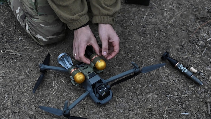 Un drone posé au sol, à l'envers. On voit les deux mains d'un soldat, à genoux, qui attachent de petites bombes dorées sous le drone.