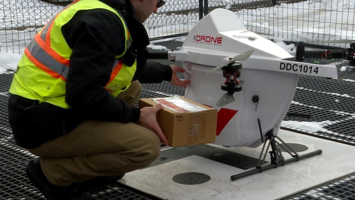 Un pilote de contrôle de drone tient un paquet en carton devant un drone posé sur le sol.