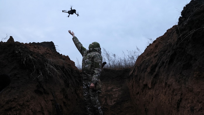 Un soldat dans une tranchée s'apprête à reprendre dans ses mains un drone. 