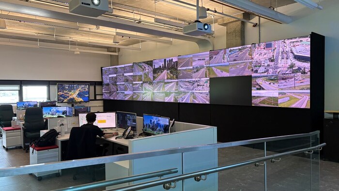 Un homme est assis devant un ordinateur dans un centre de contrôle. Un gros écran de télévision qui projette des images du trafic se trouve devant lui.