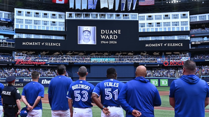 Les joueurs et les entraîneurs regardent un écran géant montrant le nom et la photo de Duane Ward, avec les chiffres 1964-2026.
