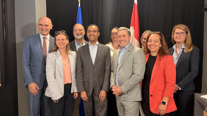 Neuf personnes posent ensemble en souriant. Derrière elles, deux drapeaux dépassent, celui du Québec à gauche et celui du Canada à droite. 