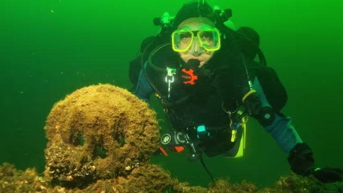 Durrell Martin sous l'eau et en équipement de plongée au bord d'une épave recouverte de coquillages.