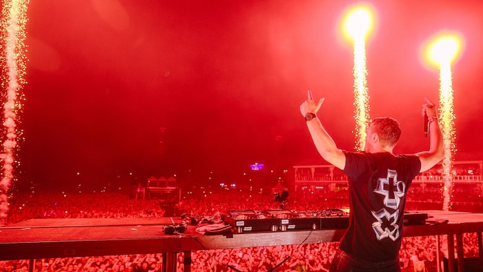 Un DJ lève les bras devant sa platine et une foule immense rassemblée pour un concert avec pyrotechnie.