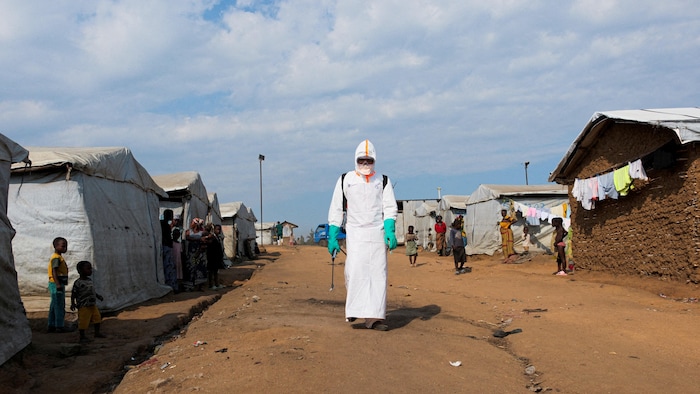 Un homme vêtu d'une combinaison blanche marche sur un sol en terre entourée de tentes près desquelles se tiennent des femmes et des enfants.
