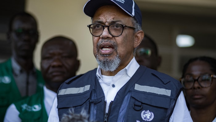 Tedros Adhanom Ghebreyesus, portant une casquette et une veste de l'OMS, s'exprime devant des micros.