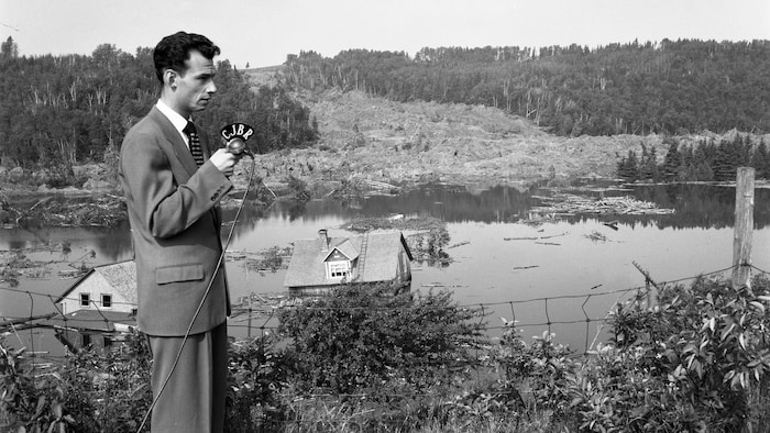 Un journaliste en complet tient un microphone de CJBR. On aperçoit derrière lui deux maisons poussées dans la rivière par les éboulis.