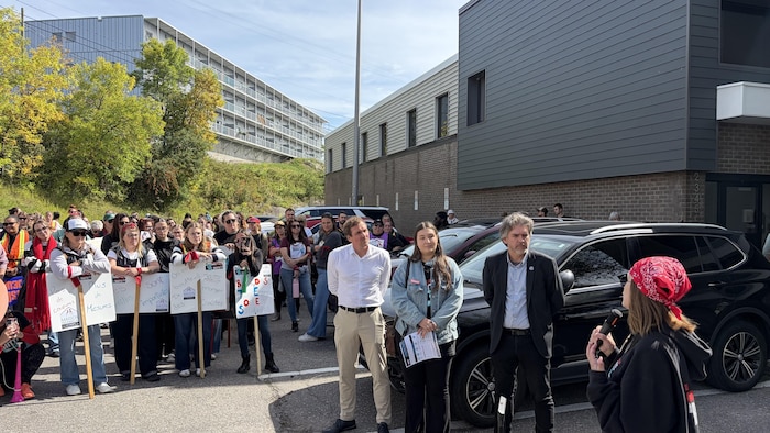 Les manifestants et les candidats écoutent une manifestante qui a un micro en main.