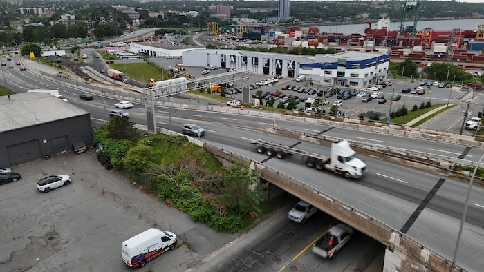 Un semi-remorque passe sur une autoroute qui enjambe une rue étroite. Un grand entrepôt et un terminal de conteneurs apparaissent au loin.