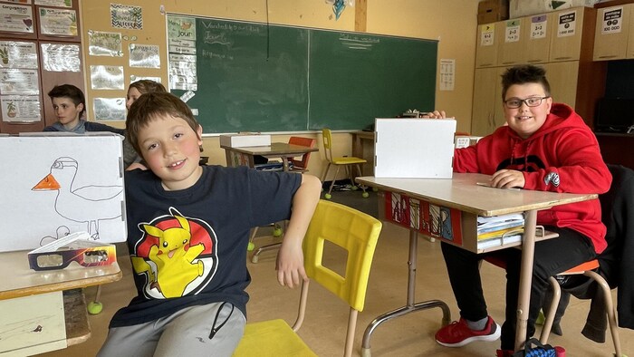 Édouard Lacharité Gourd et Arthur Cadieux posent avec leurs boîtes à éclipse assis à leur pupitre en classe.
